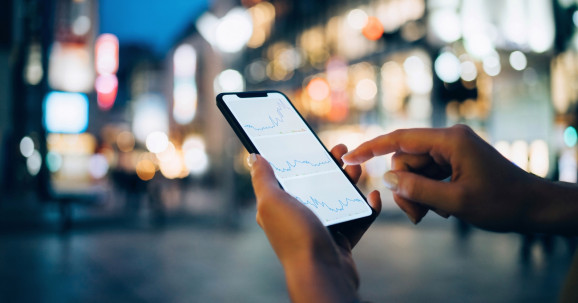 Businesswoman reading financial trading data on smartphone in city street against illuminated urban skyscrapers